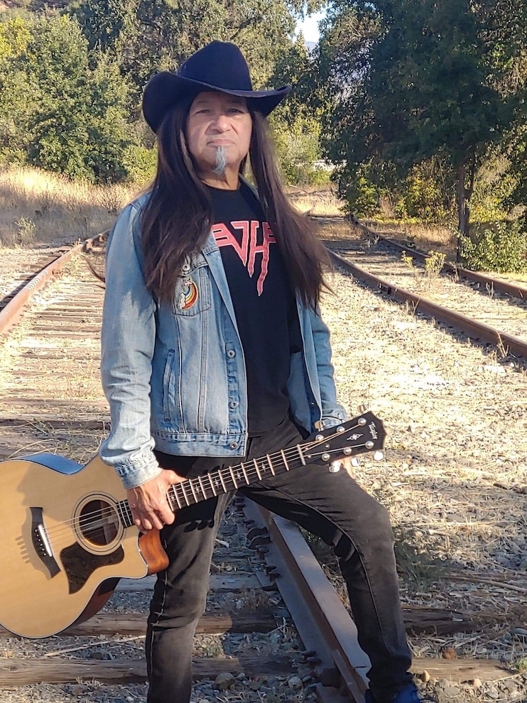 Musician Raven Blackwing with guitar standing on railroad tracks