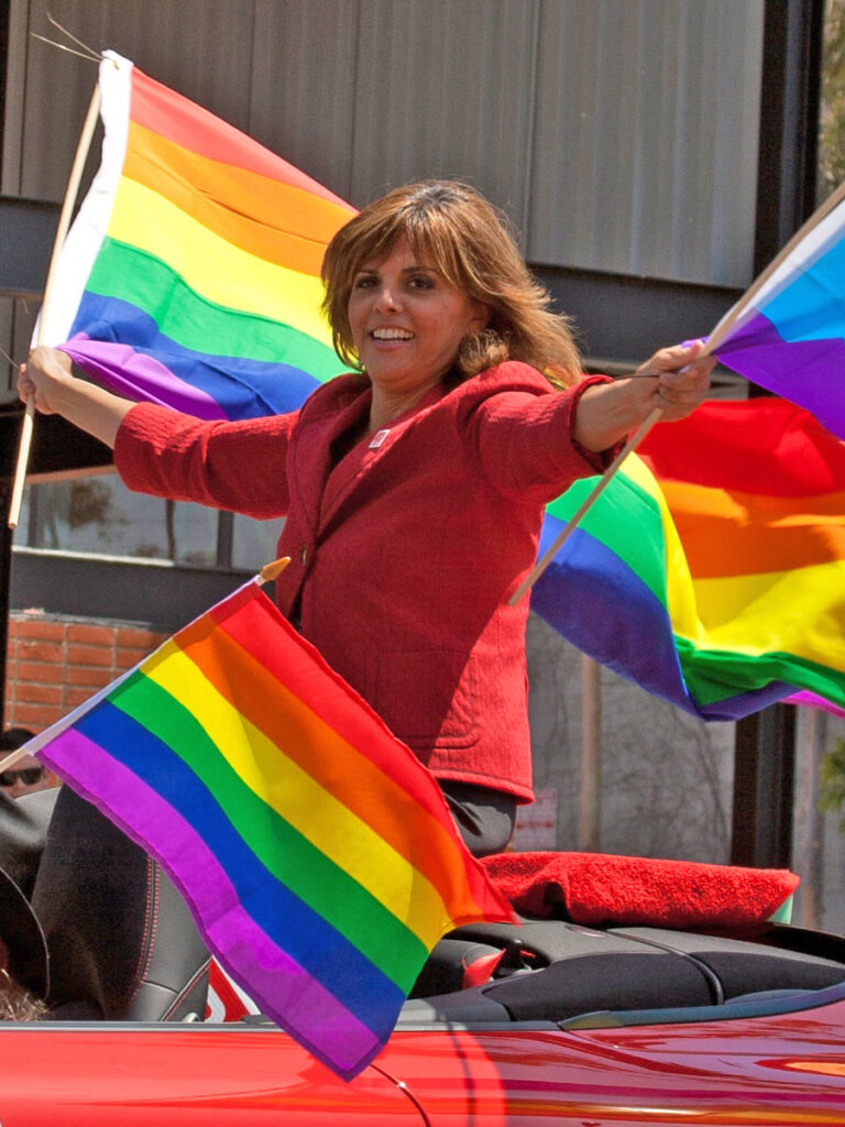 Gay vegan journalist Jane Velez-Mitchell waves three Pride flags while riding in a car